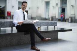 Positive young male entrepreneur in elegant formal outfit sitting in railway station waiting hall holding newspaper, cheerful economist reading daily press satisfied with financial news during break