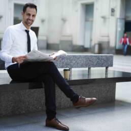 Positive young male entrepreneur in elegant formal outfit sitting in railway station waiting hall holding newspaper, cheerful economist reading daily press satisfied with financial news during break