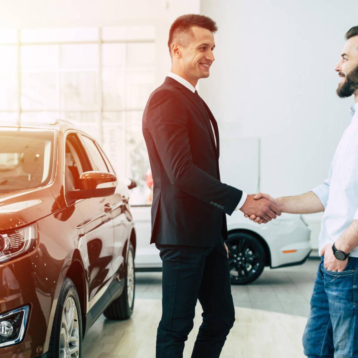 The manager in the dealership shakes hands with a young man in a light shirt against a new car.