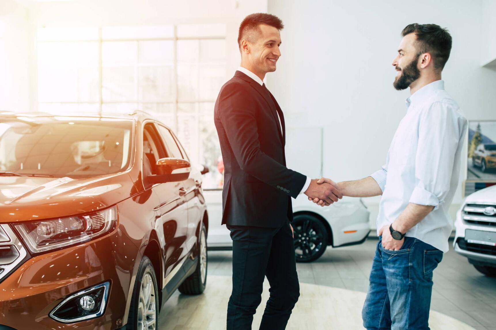 The manager in the dealership shakes hands with a young man in a light shirt against a new car.