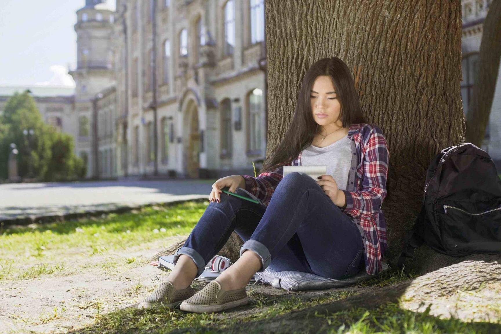 International student sitting under tree, holding notebook, looking at schedule