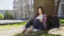 International student sitting under tree, holding notebook, looking at schedule