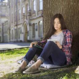 International student sitting under tree, holding notebook, looking at schedule
